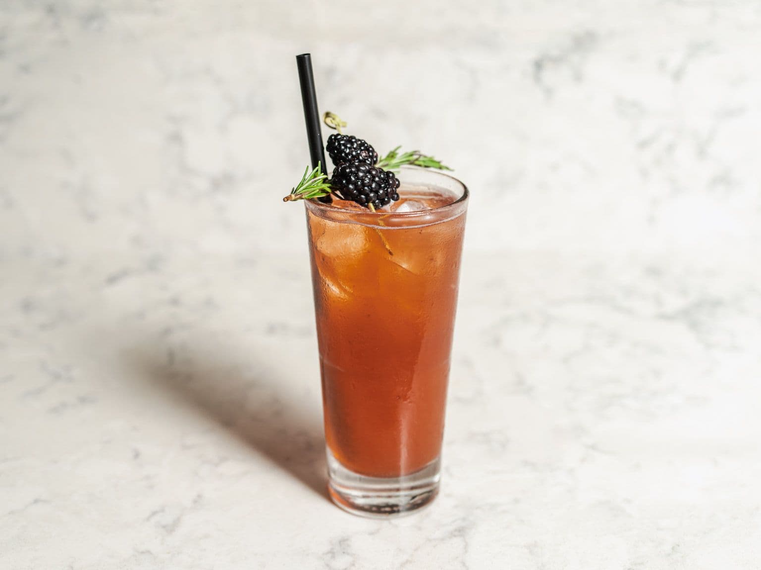 A tall iced cocktail garnished with a blackberry and a sprig of rosemary.