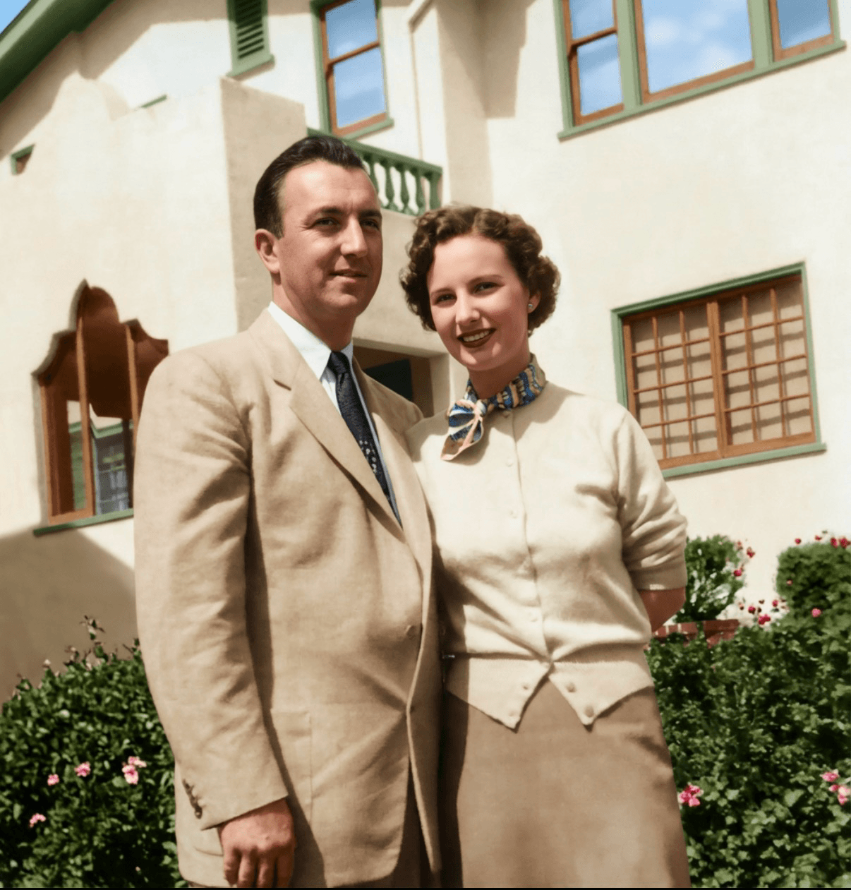 Colorized photo of Jack Holder in a light suit and tie standing beside his wife, who wears a cream cardigan and a patterned scarf, in front of a stucco house with flowering shrubs.