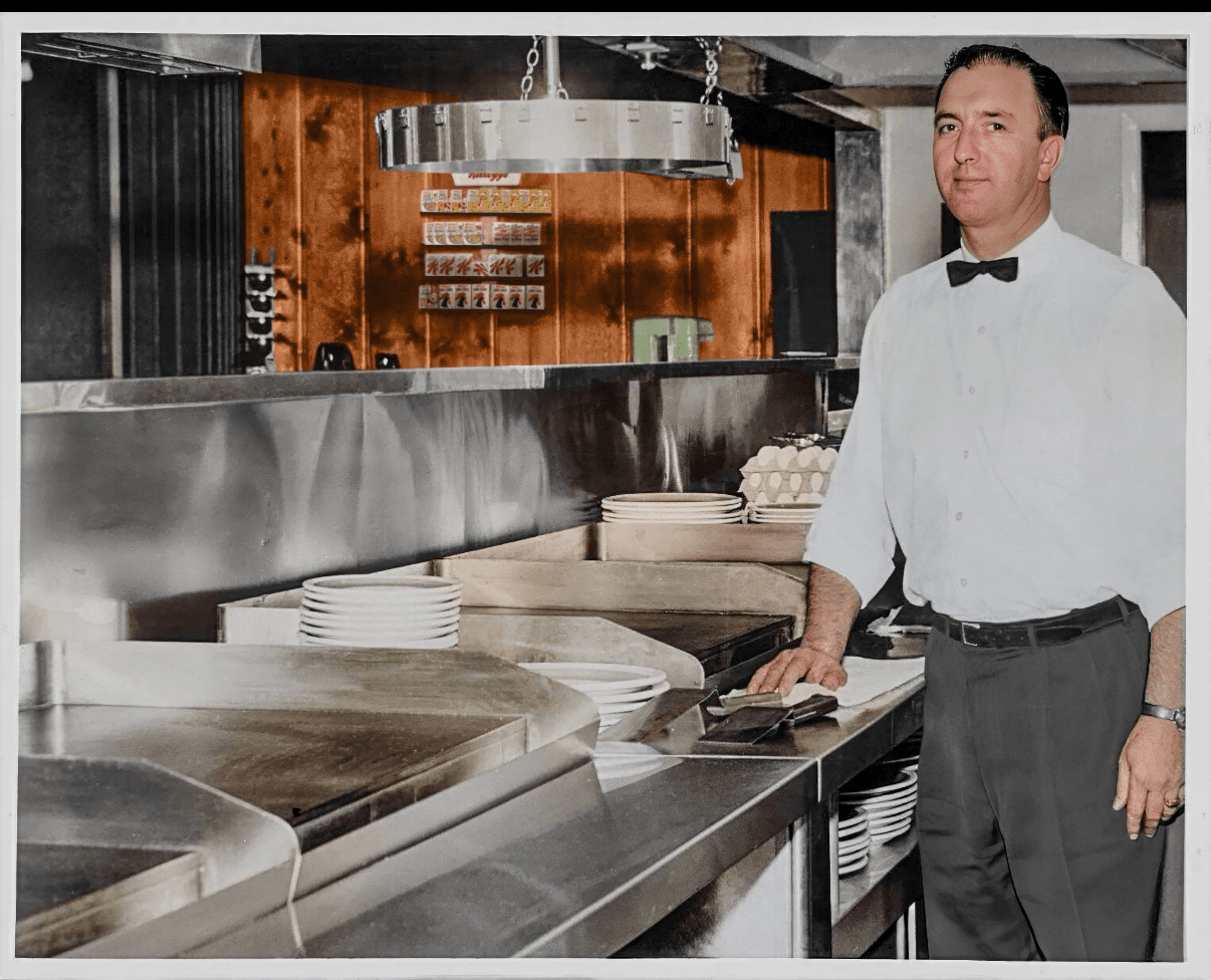 Colorized photo of Jack Holder in a white shirt and black bow tie, standing at a stainless steel griddle line with stacks of plates and wood-paneled walls behind him.