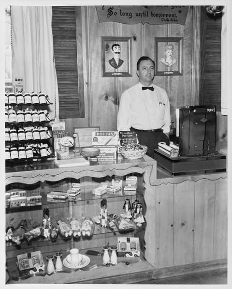 Black-and-white photo of Jack Holder in a white shirt and bow tie behind a wood-paneled counter with a cash register, candy and souvenir displays. A sign above him reads “So long until tomorrow.”