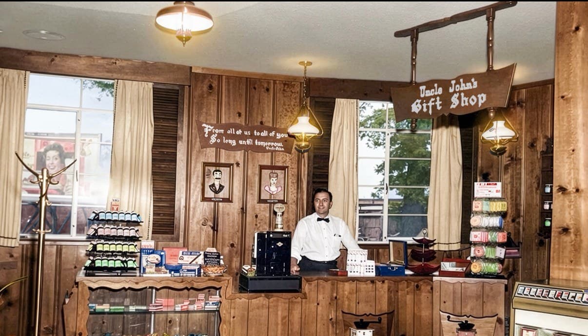 Colorized photo of a wood-paneled gift shop with a hanging sign reading Uncle John's Gift Shop. A man in a bow tie stands behind the counter among candy and souvenir displays.