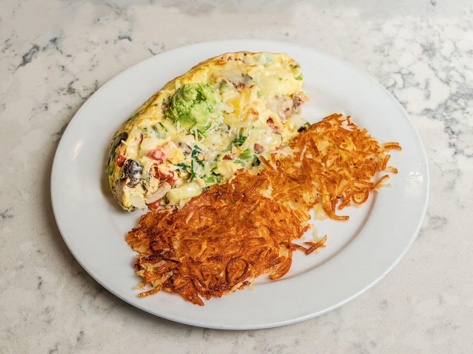 A folded omelette with avocado and vegetables beside golden hash browns.