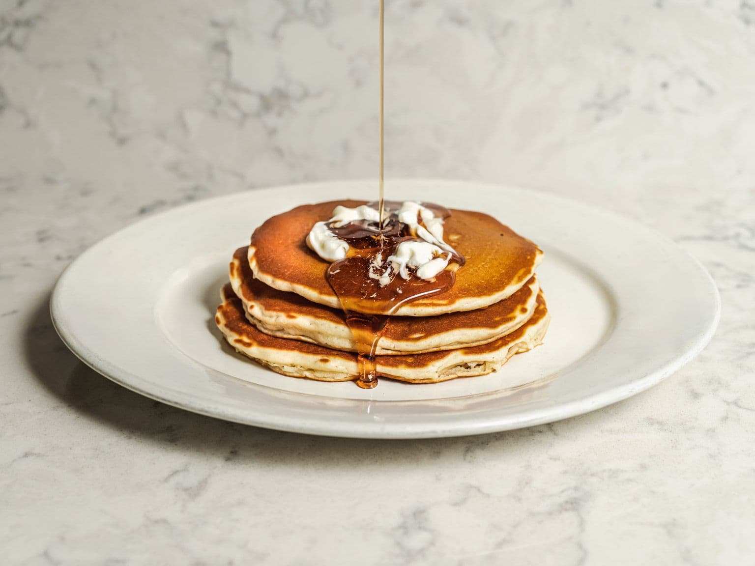 Syrup pouring onto a stack of three buttermilk pancakes topped with butter.