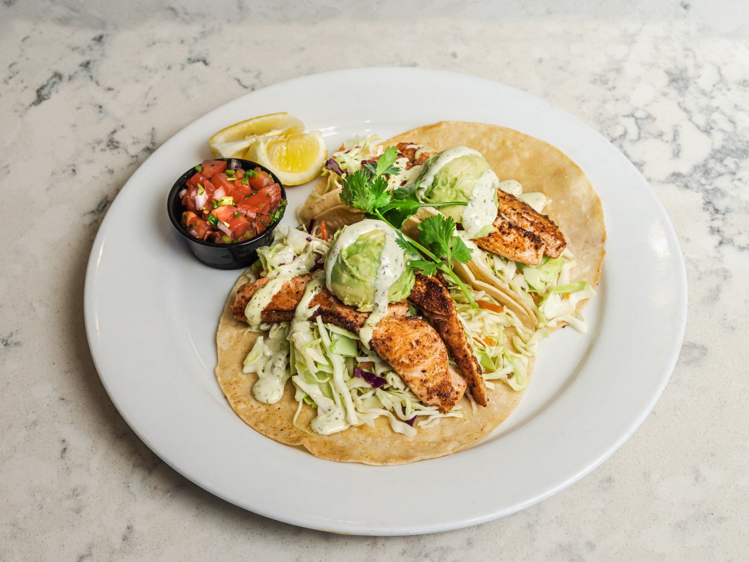 Chicken tacos with salsa, avocado, soft tortillas, lemon wedges, and sour cream on a white plate.