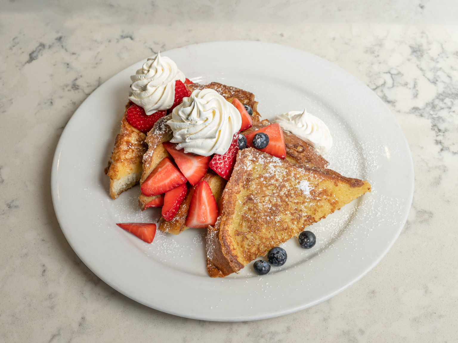 French toast with strawberries, blueberries, whipped cream on top on a white plate.