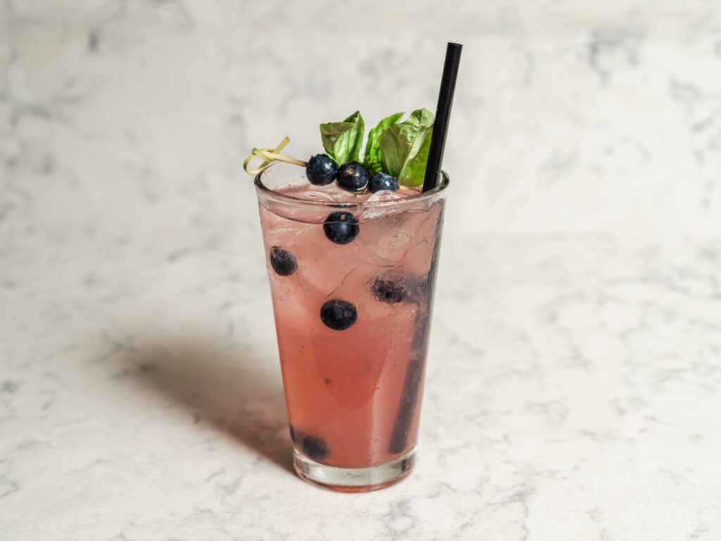 Pink cocktail with blueberries and basil on rim in a tall glass with a straw.